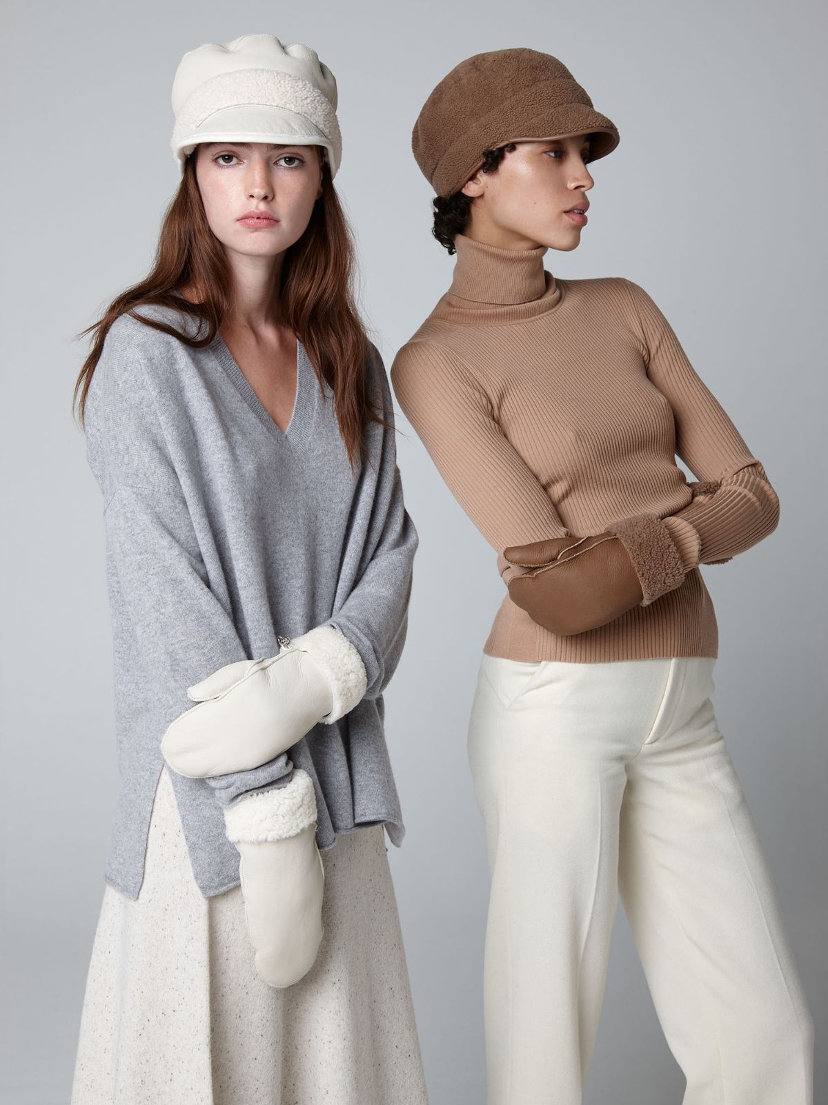 woman on grey background wearing white full palm shearling mittens next to woman wearing camel pair of full palm shearling mittens by Gushlow & Cole. both wearing a shearling cap