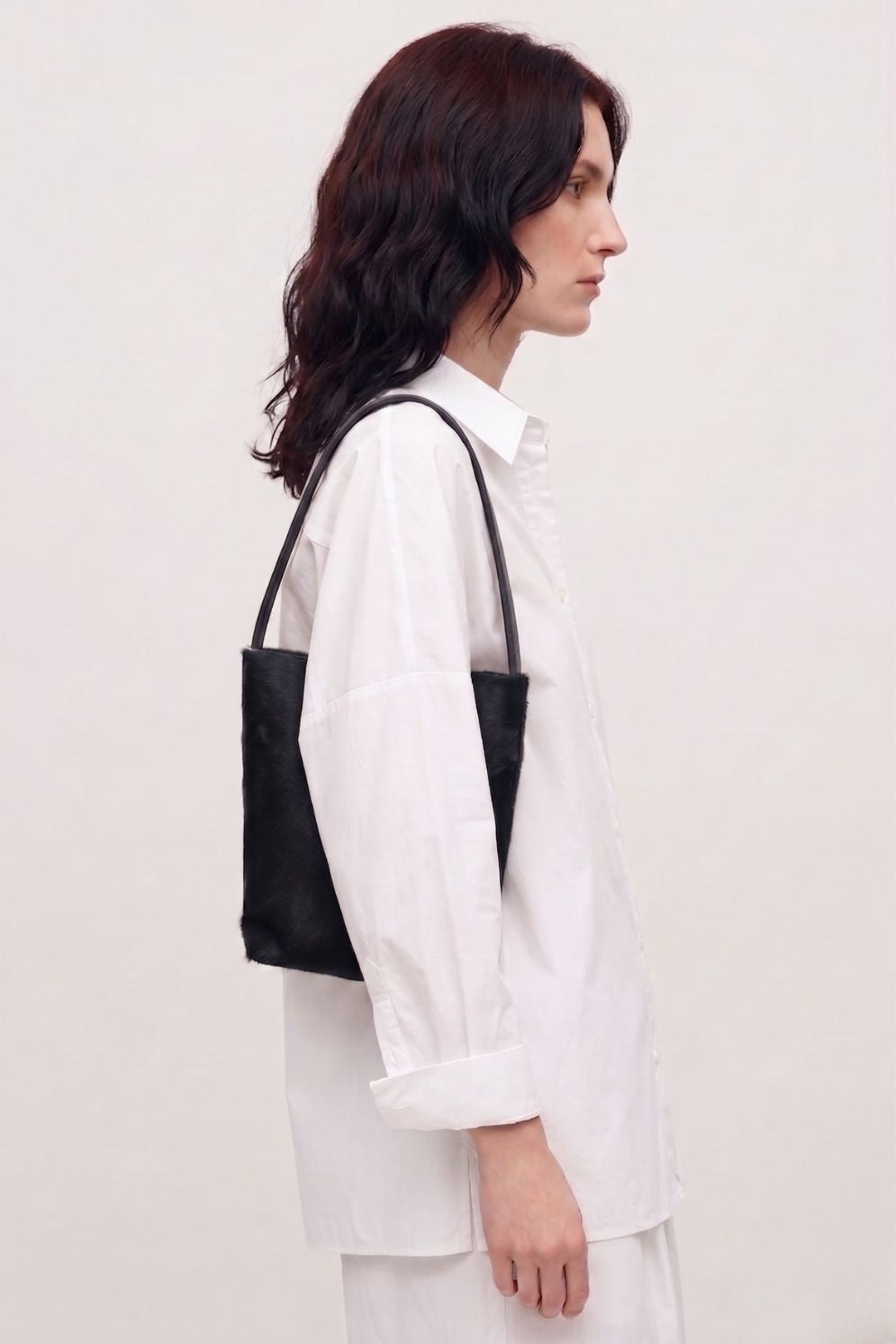 woman wearing white shirt and maye small black bucket tote bag by gushlow & cole on white background