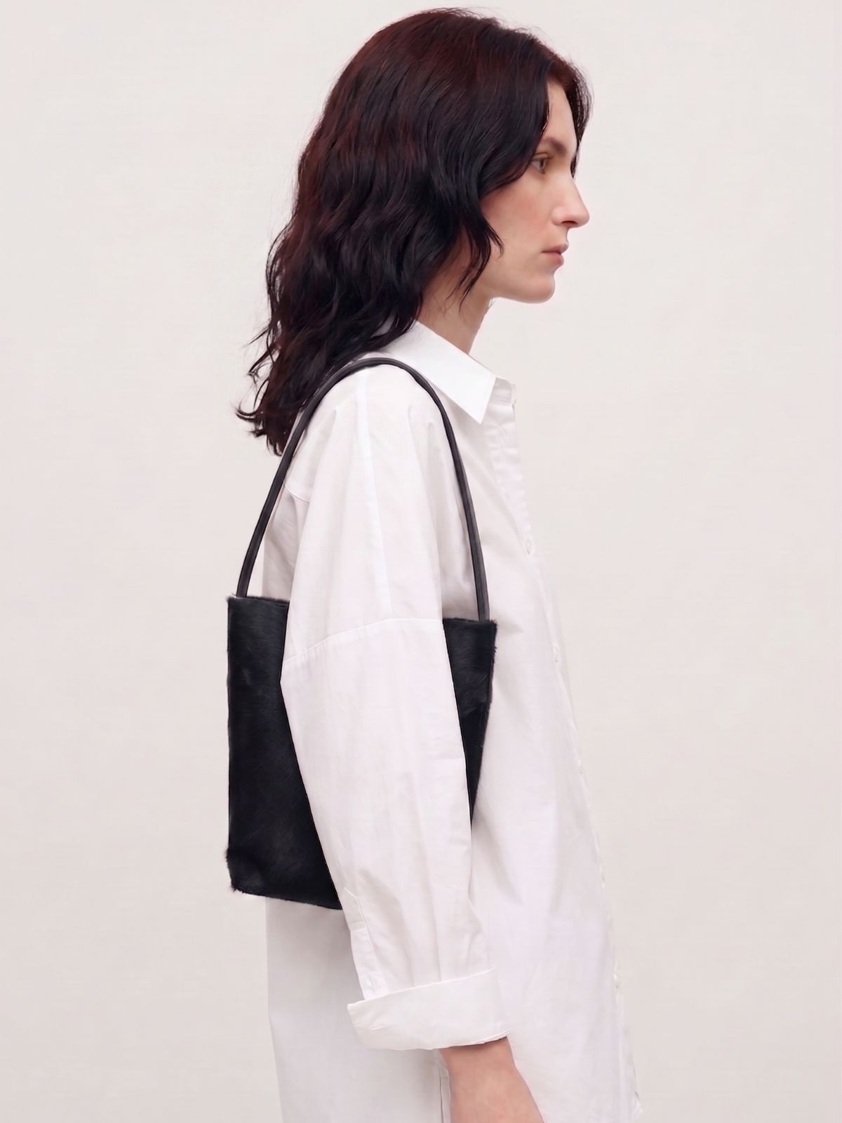 woman wearing white shirt and maye small black bucket tote bag by gushlow & cole on white background
